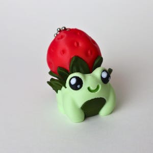 May include: A novelty keychain depicting a light green frog with large eyes and a smiling mouth, topped with a red strawberry with green leaves. The keychain is attached with a small silver chain. The frog and strawberry are likely made of plastic.
