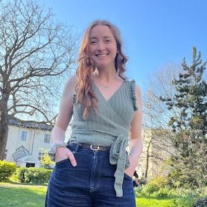 May include: A sage green crochet wrap top with a V-neck and tie closure, paired with dark wash denim jeans. The top features ruffled detailing on the shoulders. A brown belt with a gold buckle cinches the waist. A scrunchie is on the wrist.