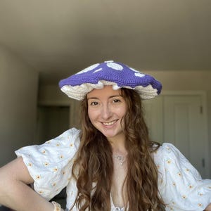 May include: A handmade crochet hat shaped like a mushroom, featuring a vibrant purple cap with white circular spots and a white ruffled underside. The hat has a wide, flat top and a textured, stitched construction throughout the entire piece.