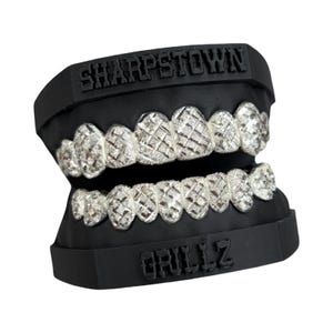 May include: A set of silver-colored grillz with a diamond-cut pattern, set in a black mold. The word "SHARPSTOWN" is embossed on the top, and "GRILLZ" is embossed on the bottom.