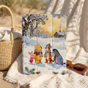 Op de afbeelding: Een tablet hoesje met een winterse scène illustratie van Winnie de Poeh, Knorretje, Tijger en Iejoor. De afbeelding toont de personages in een besneeuwd landschap met een hut en bomen. De hoes heeft een beschermhoes.