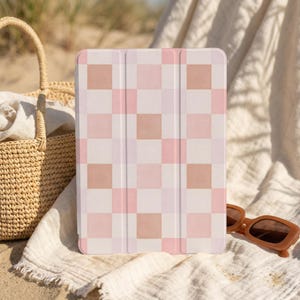 May include: A tablet case with a pink and white checkered pattern. The case is resting on a white cloth next to a woven basket and brown sunglasses. The case has a magnetic cover.