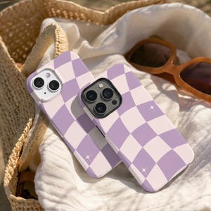 May include: Two phone cases with a light purple and white chequered pattern. The cases are on a white cloth, with a straw bag and sunglasses in the background. The cases have a glossy finish and a star design.