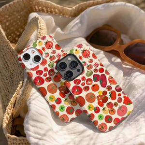 May include: Two phone cases with a repeating pattern of red, orange, and green tomatoes on a cream background. The cases are lying on a white cloth, with a straw bag and sunglasses in the background.