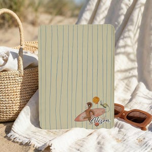 May include: A beige notebook featuring blue vertical lines and a cartoon illustration of a monkey surfing. The notebook has the name "Alison" written on it. A woven basket and sunglasses are in the background.