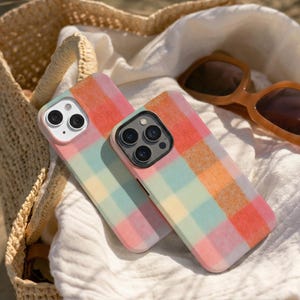 May include: Two colorful phone cases with a plaid pattern in shades of pink, orange, yellow, and blue. The cases are resting on a white cloth next to a straw bag and sunglasses.