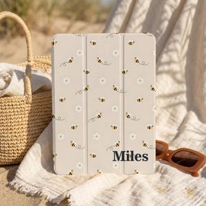 May include: Beige tablet case with a pattern of bees and white flowers. The case has the name "Miles" printed in black. The case is resting on a white cloth next to a woven basket and brown sunglasses.