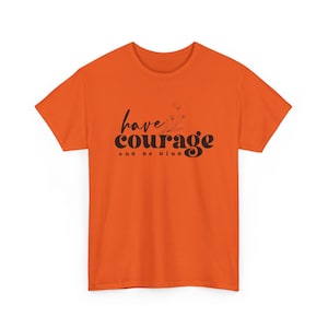 Könnte beinhalten: Orangefarbenes T-Shirt mit dem schwarzen Schriftzug "have courage and be kind". Das T-Shirt hat kurze Ärmel und einen Rundhalsausschnitt. Der Text ist mittig auf der Vorderseite des Shirts platziert.