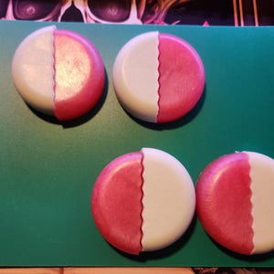 May include: Four round, half-and-half soaps. Each soap is divided into two sections: one half is white, and the other is a shimmery pink. The soaps are arranged on a green cutting board. The soaps have a wavy edge where the two colors meet.