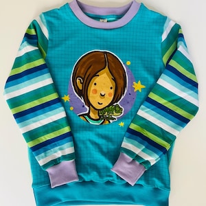 Könnte beinhalten: Ein langärmeliges Sweatshirt in Blaugrün mit Gittermuster und einer zentralen kreisförmigen Grafik einer Person mit einem grünen Chamäleon. Die Ärmel haben horizontale Streifen in Blau, Grün und Weiß, mit hellvioletten gerippten Bündchen und Kragen.