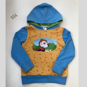 Könnte beinhalten: Ein blau-gelber Kapuzenpullover mit einer Cartoon-Grafik. Die Kapuze und die Ärmel sind blau, während der Körper gelb mit braunen Punkten ist. Auf der Vorderseite ist eine Cartoon-Figur abgebildet. Die Nummer "104" ist oben links sichtbar.