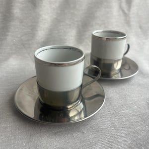 May include: Two white porcelain espresso cups with matching saucers, each featuring a polished silver-tone metal base and a thin silver rim around the top edge. The cups have a curved handle and sit securely within the metallic saucer holders.