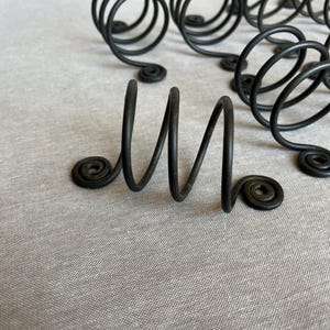 May include: Black metal napkin rings in a spiral design. The napkin rings have a matte finish and a decorative swirl at the base. The background is a light gray fabric. The napkin rings are a decorative element for table settings.