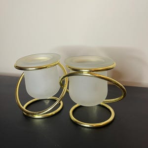 May include: Two candle holders with frosted glass cups and gold-colored metal frames. The holders have a circular base and a curved handle that wraps around the glass. The rims of the glass cups are also gold-colored.
