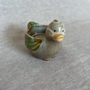 May include: A small, glazed ceramic duck-shaped bowl with a glossy finish. The duck features a muted grey body, a light orange beak, and green-toned wings. The center of the duck's back is hollowed out, creating a small, open basin for holding items.
