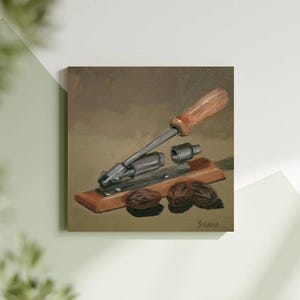 May include: A painting depicting a nutcracker with a wooden handle and metal parts, placed on a wooden base. Two nuts are shown beneath the nutcracker. The artwork is signed "SEANNA" in the bottom right corner.
