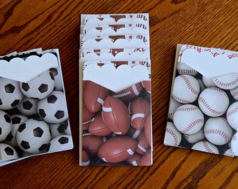 May include: Stacked gift card holders featuring soccer balls, footballs, and baseballs. Each holder has a white scalloped edge and a blank label. The holders are arranged on a wooden surface.