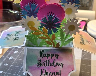 May include: A 3D pop-up greeting card featuring a bouquet of paper flowers in shades of pink, blue, white, and green. The front panel displays the text Happy Birthday Donna!, with side panels showing July 1st, 2026 and Thrive at 75 in black.