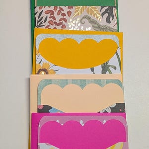 May include: A stack of colorful handmade greeting cards with scalloped edges. Each card features a different patterned paper, including floral, rainbow, and leaf designs. The cards are in shades of purple, blue, green, yellow, peach, and pink.