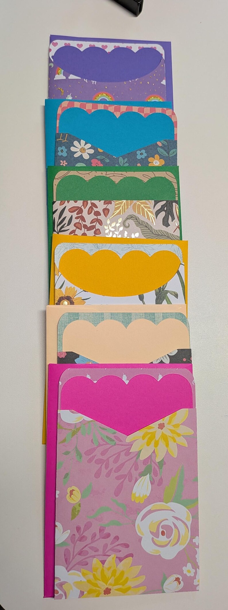 May include: A stack of colorful handmade greeting cards with scalloped edges. Each card features a different patterned paper, including floral, rainbow, and leaf designs. The cards are in shades of purple, blue, green, yellow, peach, and pink.