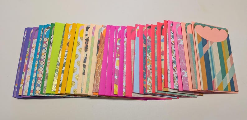 May include: A collection of colorful handmade envelopes in various patterns and colors, including purple, blue, green, yellow, pink, and patterned designs. The envelopes are arranged in a fan shape, showcasing their diverse designs.