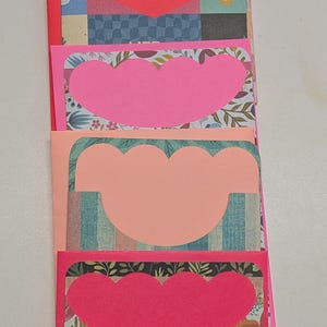 May include: A stack of handmade greeting cards with scalloped heart cutouts. The cards feature a variety of patterned papers, including floral, plaid, and geometric designs. The edges are bright pink.