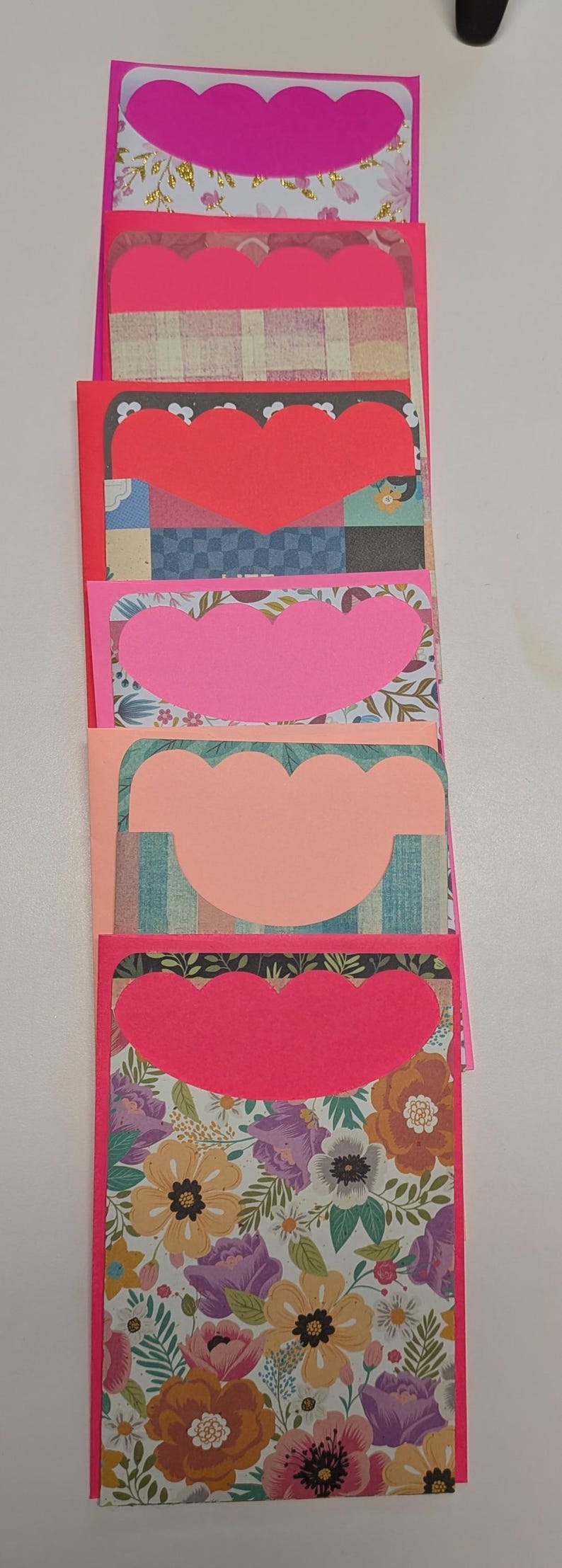May include: A stack of handmade greeting cards with scalloped heart cutouts. The cards feature a variety of patterned papers, including floral, plaid, and geometric designs. The edges are bright pink.