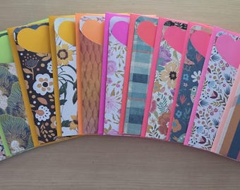 May include: A collection of colorful, patterned notebooks with heart-shaped cutouts. The notebooks feature various designs, including floral, geometric, and animal prints, with a range of colors like purple, yellow, and pink. The notebooks are arranged in a fan shape.