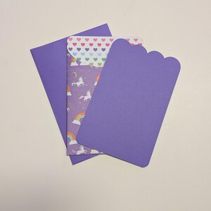 May include: A set of purple stationery items, including an envelope, a card with a scalloped edge, and a patterned insert. The insert features unicorns, rainbows, and hearts on a purple background. The envelope and card are solid purple.