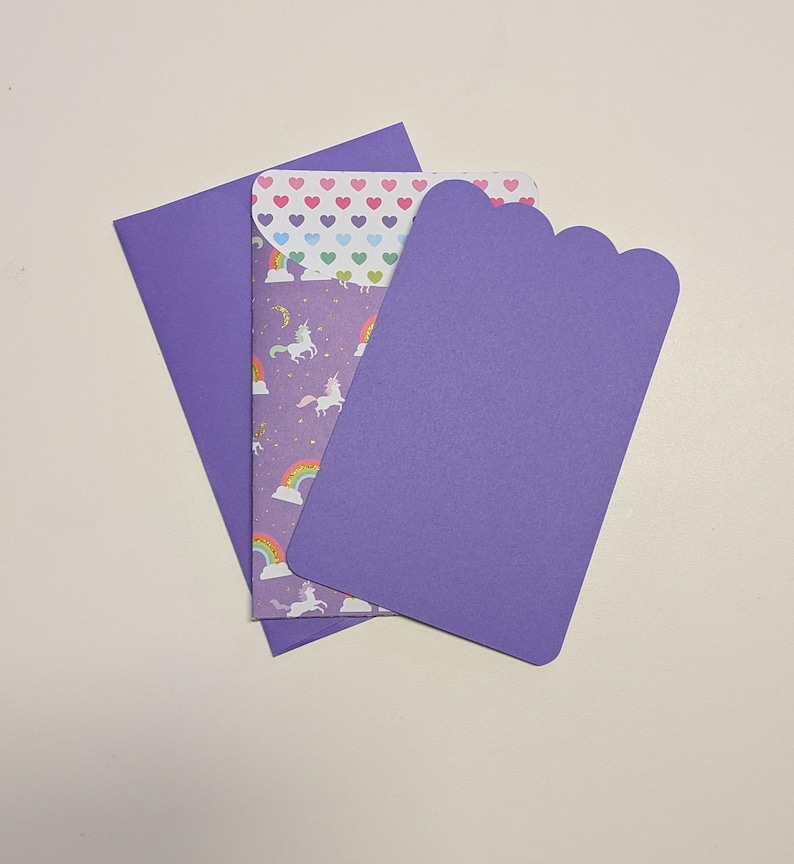 May include: A set of purple stationery items, including an envelope, a card with a scalloped edge, and a patterned insert. The insert features unicorns, rainbows, and hearts on a purple background. The envelope and card are solid purple.
