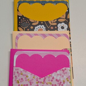 May include: A stack of colorful handmade greeting cards with scalloped edges. The cards feature various patterned papers, including floral, geometric, and confetti designs. The color palette includes purple, blue, green, yellow, peach, and pink.