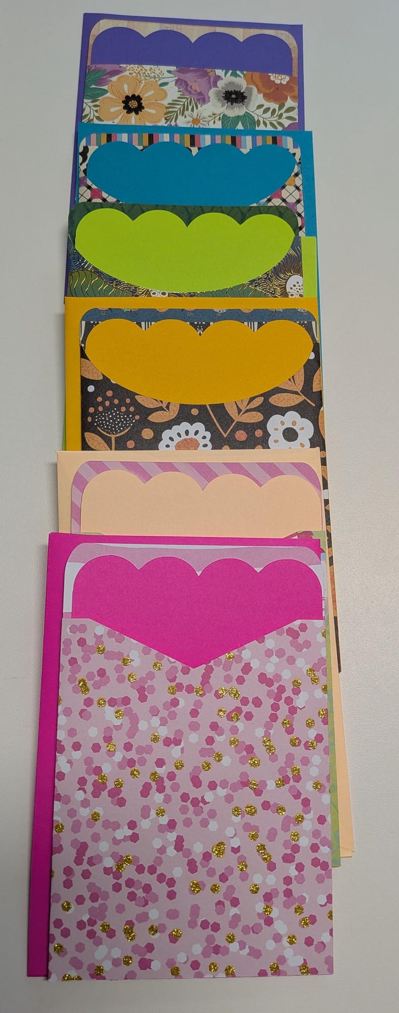 May include: A stack of colorful handmade greeting cards with scalloped edges. The cards feature various patterned papers, including floral, geometric, and confetti designs. The color palette includes purple, blue, green, yellow, peach, and pink.