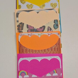 May include: A stack of colorful handmade paper pockets with scalloped heart-shaped cutouts. The pockets feature various patterned papers with designs including butterflies, gold hearts, rainbows, and unicorns. The color palette includes purple, blue, yellow, orange, and pink.