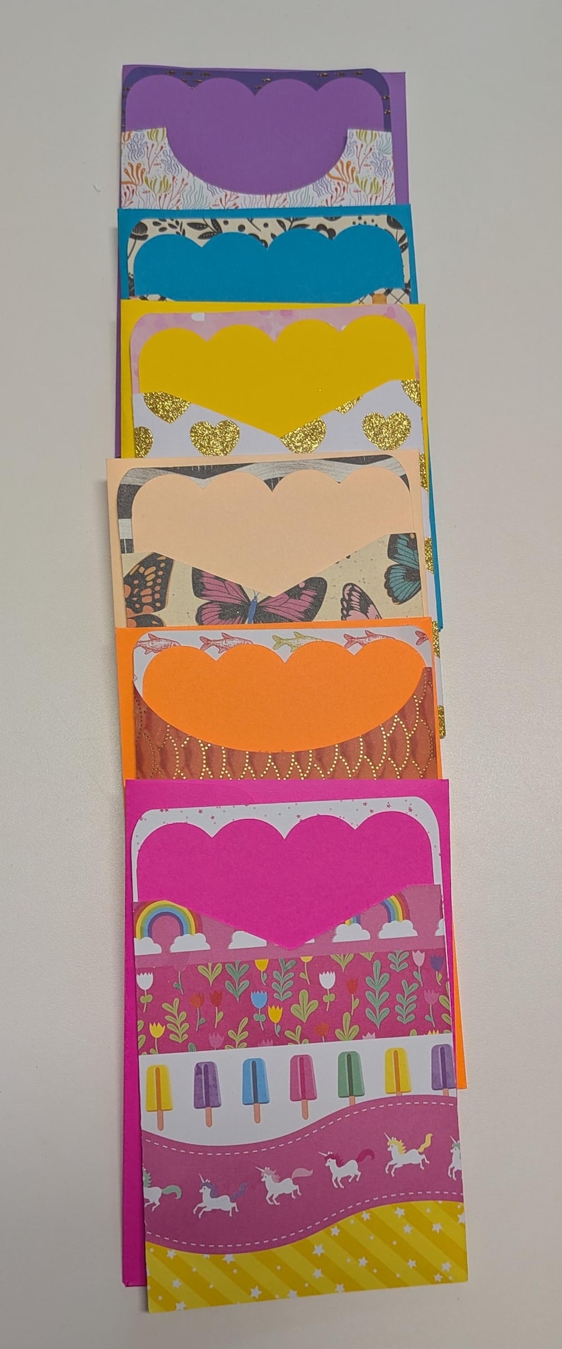May include: A stack of colorful handmade paper pockets with scalloped heart-shaped cutouts. The pockets feature various patterned papers with designs including butterflies, gold hearts, rainbows, and unicorns. The color palette includes purple, blue, yellow, orange, and pink.