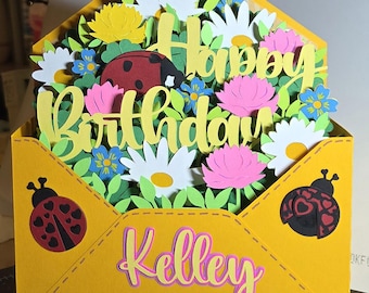 May include: A yellow paper envelope card featuring the text Happy Birthday in yellow script and the name Kelley in pink and yellow. The card is filled with layered paper cutouts of white daisies, pink flowers, and a red ladybug with black spots.