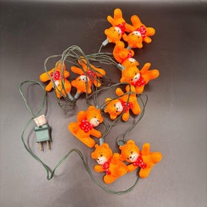 May include: A string of orange teddy bear Christmas lights. Each bear has a red scarf and a smiling face. The lights are connected by green wire and have a green plug.