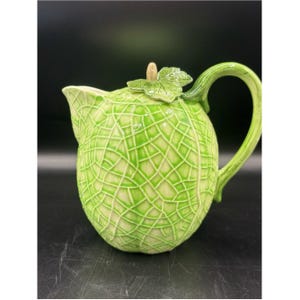 May include: A ceramic teapot shaped like a cantaloupe melon with a light green and cream textured rind pattern. The lid features a sculpted green leaf and a small stem handle. It has a curved green handle and a pointed spout on the side.