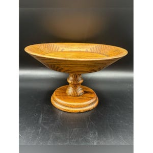 Puede incluir: Un cuenco de madera de color marrón medio con un pedestal que presenta una base ancha y poco profunda con un patrón de veta de madera visible. El cuenco está sostenido por un tallo central torneado que se conecta a una base circular plana.