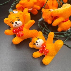May include: A string of orange teddy bear Christmas lights. Each bear has a white face, black eyes, and a red bow with white polka dots. The lights are strung on a green wire.
