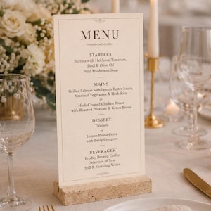 Può includere: Un menu stampato con la parola "MENU" in alto, che elenca antipasti, piatti principali, dessert e bevande. Il menu è esposto su una base di pietra colorata, con candele e composizioni floreali sullo sfondo.