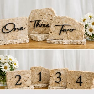 Pode incluir: Sinais de números de mesa de pedra com letras pretas. Os números incluem One, Two, Three e os números 1, 2, 3 e 4. Os sinais são bege e têm uma textura áspera e natural. Margaridas brancas estão no fundo.