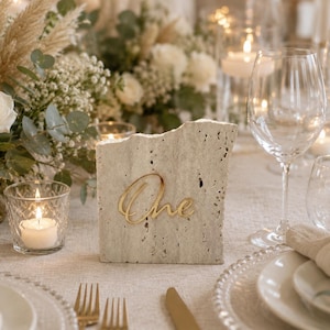 Puede incluir: Decoración de mesa para una boda con un número de mesa de hormigón con la palabra "One" en escritura dorada. La mesa está puesta con platos blancos, cubiertos dorados y copas de vino. Una vela encendida está en un portavelas de cristal.