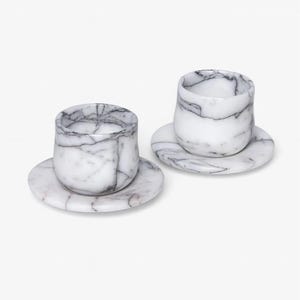 May include: Two marble espresso cups and saucers. The cups are cylindrical with rounded bottoms, and the saucers are round. The marble has a white base with grey veining. The set is displayed on a white background.
