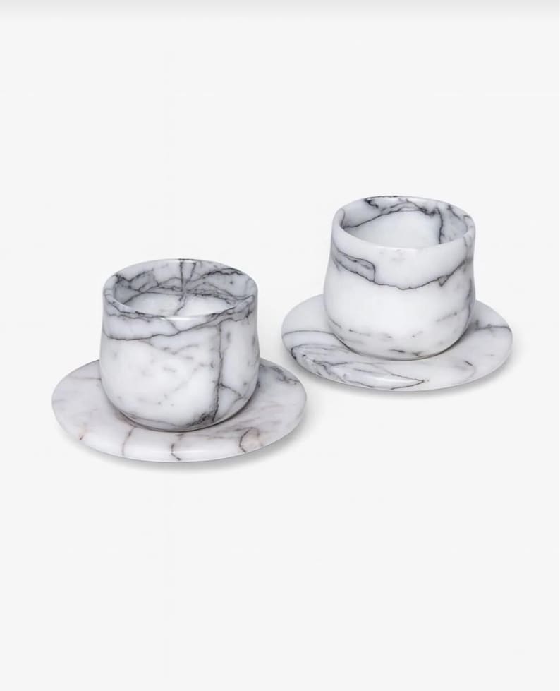 May include: Two marble espresso cups and saucers. The cups are cylindrical with rounded bottoms, and the saucers are round. The marble has a white base with grey veining. The set is displayed on a white background.