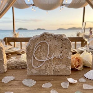 May include: A rough-hewn, beige travertine stone table number sign featuring the white script text "One" centered on the front. The stone has a porous, natural texture and is displayed on a wooden surface alongside scattered white seashells.