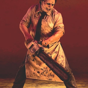 May include: A figure wearing a mask made of skin, a light-colored collared shirt, a dark tie, and a stained apron. The figure holds a large chainsaw with both hands and wears dark trousers and brown shoes, standing in a wide stance.