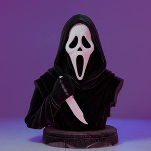 May include: A black bust of the Ghostface killer from the Scream horror film series. The figure wears a black hooded robe and a white mask with black eye and mouth openings. It holds a white knife in its gloved hand. The bust sits on a dark, textured base.