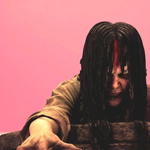 Può includere: Immagine a tema horror con una figura dalla pelle pallida con lunghi capelli scuri e bagnati che oscurano parzialmente il viso. Gli occhi della figura sono visibili e la mano si protende. Lo sfondo è rosa.