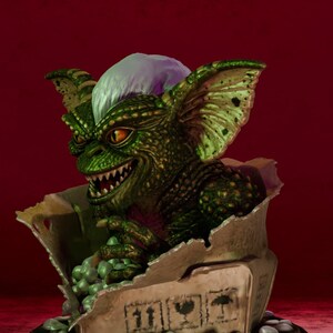 Puede incluir: Una figura de criatura verde texturizada con orejas grandes, dientes afilados y un mech&oacute;n de pelo blanco, emergiendo de una caja de cart&oacute;n rota llena de peque&ntilde;as esferas verdes. La base tiene una placa dorada con el texto Nostalgiahorror3d.