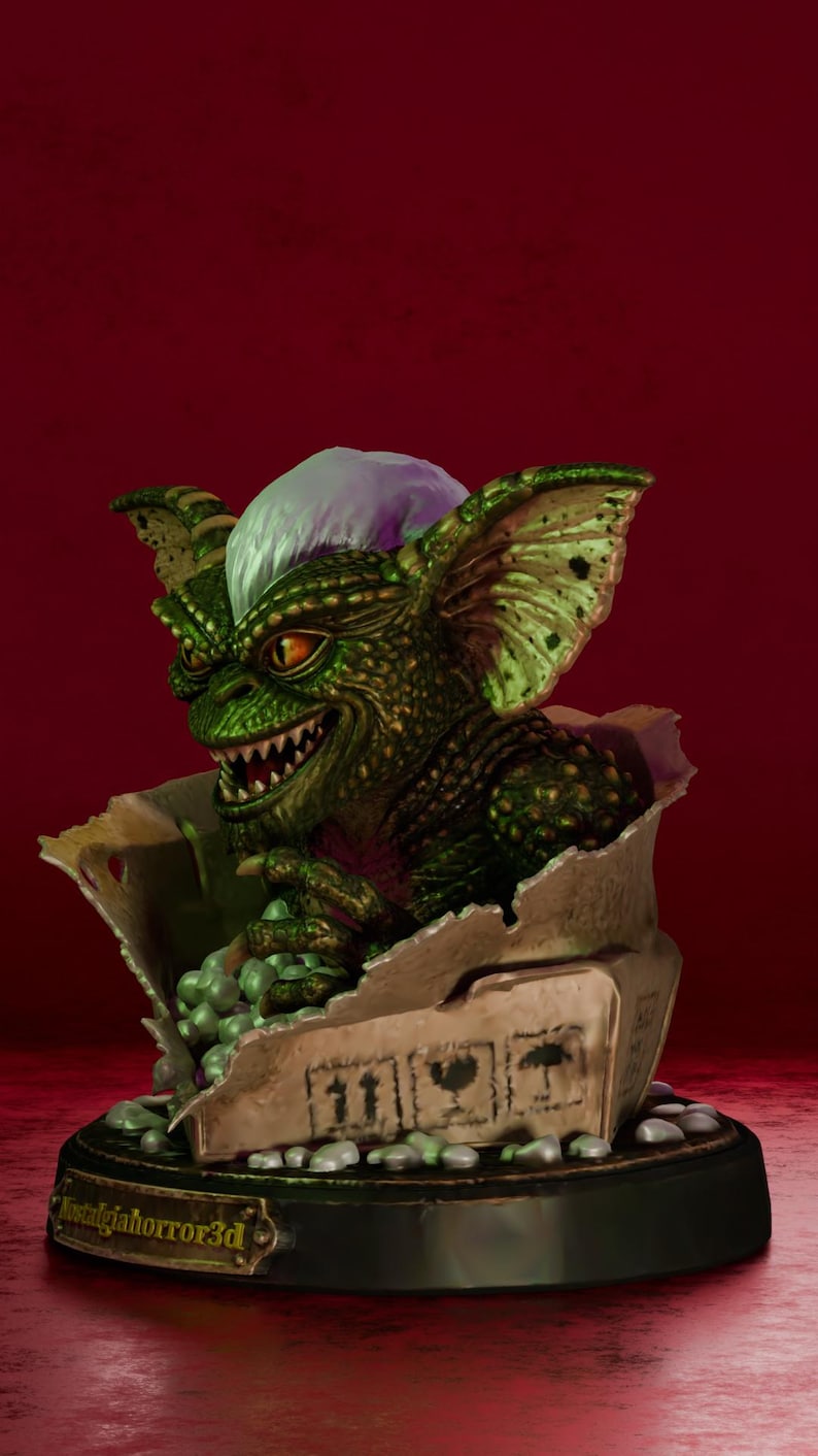 Puede incluir: Una figura de criatura verde texturizada con orejas grandes, dientes afilados y un mech&oacute;n de pelo blanco, emergiendo de una caja de cart&oacute;n rota llena de peque&ntilde;as esferas verdes. La base tiene una placa dorada con el texto Nostalgiahorror3d.